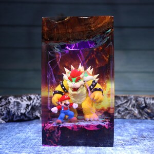Pu&ograve; includere: Un blocco rettangolare in resina contenente le statuette di Bowser e Mario. La resina trasparente presenta uno sfondo infuocato arancione e viola con effetti di fumo vorticosi, sormontato da un bordo in legno naturale scuro.