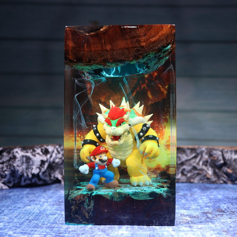 Pu&ograve; includere: Un blocco rettangolare in resina contenente una statuetta di Bowser e una di Mario. La resina presenta una parte superiore in legno scuro e un corpo traslucido con motivi a fumo blu e arancione, creando un effetto cavernoso e infuocato.