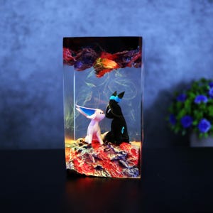 May include: A rectangular resin art piece featuring two stylized figures, one white and one black, set on a red and blue base. The figures are enclosed in a clear resin block with colorful, cloud-like details at the top.