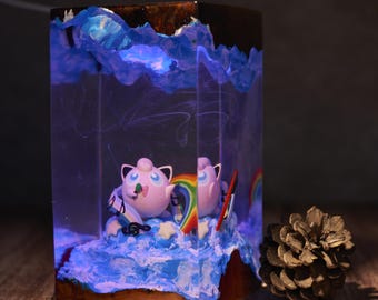 May include: A rectangular resin art piece featuring two pink cartoon characters with rainbow accents, set against a blue and white wave design. The top has a dark brown and blue design, with a pinecone on the side.