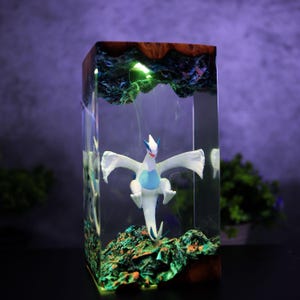 May include: A rectangular resin art piece featuring a white and blue mythical creature figurine. The figurine is encased in clear resin with a colorful, textured base and top. The piece has a wooden top and a green light.