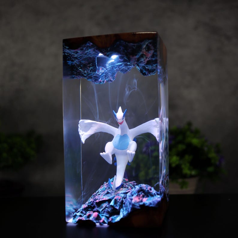 May include: A resin art block with a white and blue mythical creature figurine inside. The clear resin is encased in a dark wood frame, with a blue and purple base. The piece is lit with blue light, creating a striking display.
