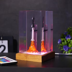 Peut inclure: Lampe décorative avec deux fusées miniatures en lancement, enfermées dans un bloc acrylique transparent. Les fusées ont des corps blancs et noirs avec des flammes orange. La base est un bloc de bois, et la lampe émet une lueur colorée.
