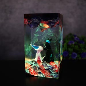 May include: A rectangular resin art piece featuring two stylized figures, one white and one black, set on a red and blue textured base. The figures are enclosed within a clear resin block with swirling green and blue accents.