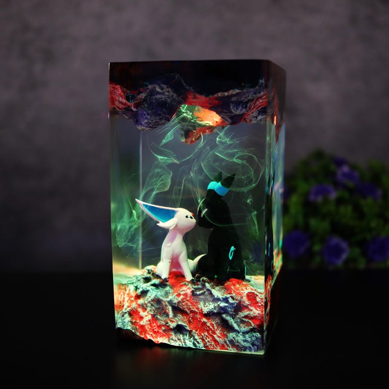 May include: A rectangular resin art piece featuring two stylized figures, one white and one black, set on a red and blue textured base. The figures are enclosed within a clear resin block with swirling green and blue accents.