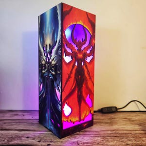 May include: A rectangular lamp featuring detailed fantasy art. One side depicts a blue and white angel figure, while the other showcases a red demon. The lamp emits a purple and pink glow, creating a dramatic effect. The lamp is plugged in and ready to illuminate.