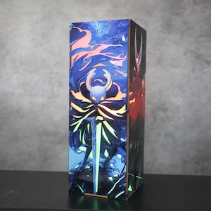 Puede incluir: Una caja rectangular con una ilustración vibrante con una combinación de colores azul, naranja y verde. La obra de arte representa un personaje con una espada brillante y un fondo detallado. La caja está de pie sobre una superficie negra.