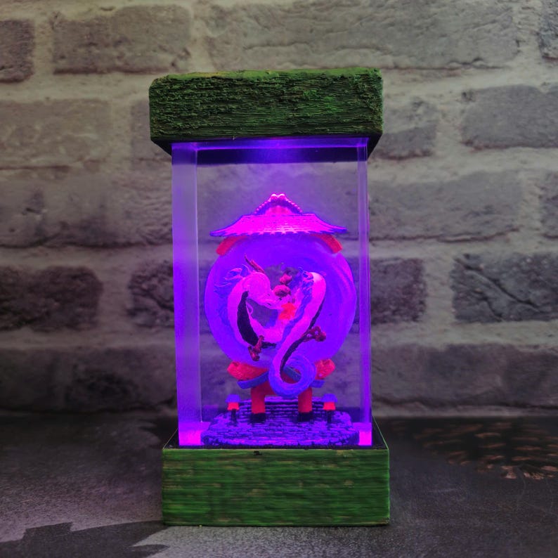 Op de afbeelding: Een decoratieve lamp met een drakenontwerp in een heldere rechthoekige behuizing. De lamp heeft een groene houten basis en bovenkant, met een paars licht dat de draak en zijn omgeving verlicht. De draak is wit en rood.