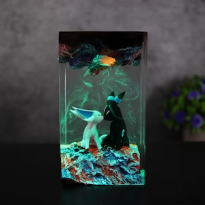 May include: A rectangular resin art piece featuring two stylized figures, one white and one black, on a colorful base. The figures are set within a clear resin block with green and orange accents, creating a glowing effect. The overall theme is fantasy.