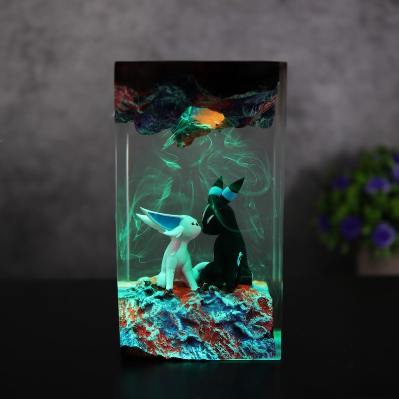 May include: A rectangular resin art piece featuring two stylized figures, one white and one black, on a colorful base. The figures are set within a clear resin block with green and orange accents, creating a glowing effect. The overall theme is fantasy.