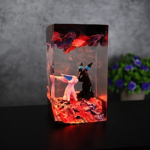 May include: A rectangular resin art piece featuring two cartoon figures, one pink and one black, in an embrace. The figures are set on a red, textured base, with red and orange lighting effects. The top of the piece has a dark, cloud-like design.