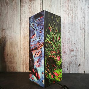 Puede incluir: Una lámpara rectangular con marco negro y dos lados con diseños coloridos e intrincados. Un lado tiene un diseño azul y rojo, mientras que el otro tiene un diseño verde y rojo. La lámpara está enchufada e iluminada.