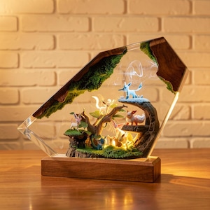 May include: A decorative lamp with a diorama of Pokémon characters in a forest scene. The scene is encased in a clear, angled resin structure with a wooden base. The diorama includes a tree, moss, and a soft, glowing light.
