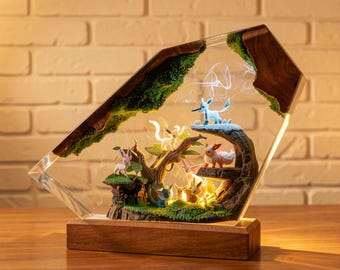 May include: A decorative lamp featuring a diorama of various Pokémon characters in a forest setting. The scene is encased in a clear, angled resin structure with a wooden base. The diorama includes a tree, moss, and a soft, glowing light.