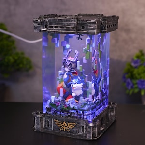 Pode incluir: Um expositor decorativo iluminado com duas personagens de desenhos animados dentro de uma caixa retangular transparente. A caixa é emoldurada por uma estrutura escura em forma de castelo. As personagens são azuis, brancas e vermelhas, e o expositor emite um brilho azul.