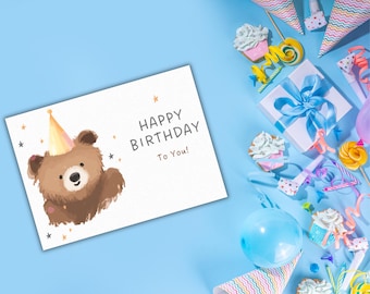 May include: A birthday card with a watercolor illustration of a brown bear wearing a party hat. The card reads "Happy Birthday To You!" with star accents. Party supplies, including cupcakes, a gift box, and balloons, are scattered around the card on a blue background.