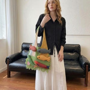 May include: A colorful, knitted tote bag featuring multiple fish appliqués. The bag has a green base with a fringe, and the fish are in shades of red, orange, gray, and green. The bag is held by a person wearing a black shirt and white skirt.