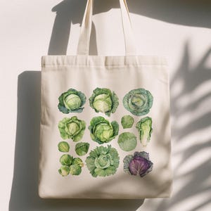 Könnte beinhalten: Eine cremefarbene Tragetasche mit einer Aquarellillustration verschiedener Kohlsorten. Das Design umfasst grüne und lila Kohlsorten, Rosenkohl und Kohlblätter. Die Tasche hat zwei Griffe und ist aus einem robusten Stoff gefertigt.