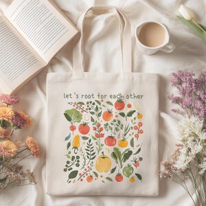 Puede incluir: Bolso tote color crema con una colorida ilustración de verduras y el texto "let's root for each other." El diseño incluye tomates, pimientos y verduras de hoja verde. El bolso tiene dos asas y está sobre una superficie blanca.
