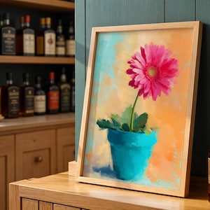 Op de afbeelding: Een ingelijste kunstdruk met een roze gerbera in een turquoise pot. Het kunstwerk staat tegen een oranje en blauwe achtergrond en is gepresenteerd in een licht houten frame.