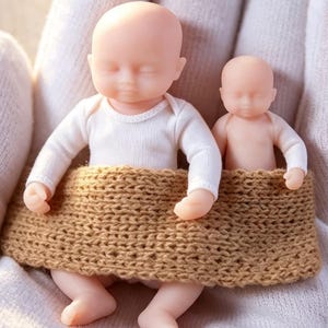 May include: Two miniature baby figurines with smooth, pale skin tones. The larger figurine wears a long-sleeved white shirt, while the smaller one is bare-chested. Both are tucked into a single, textured, tan-colored knitted fabric wrap.