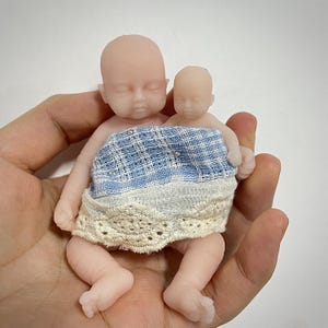 May include: A pair of miniature baby dolls with closed eyes. The dolls are wearing a blue and white patterned skirt with a lace trim. The dolls are held in a hand.