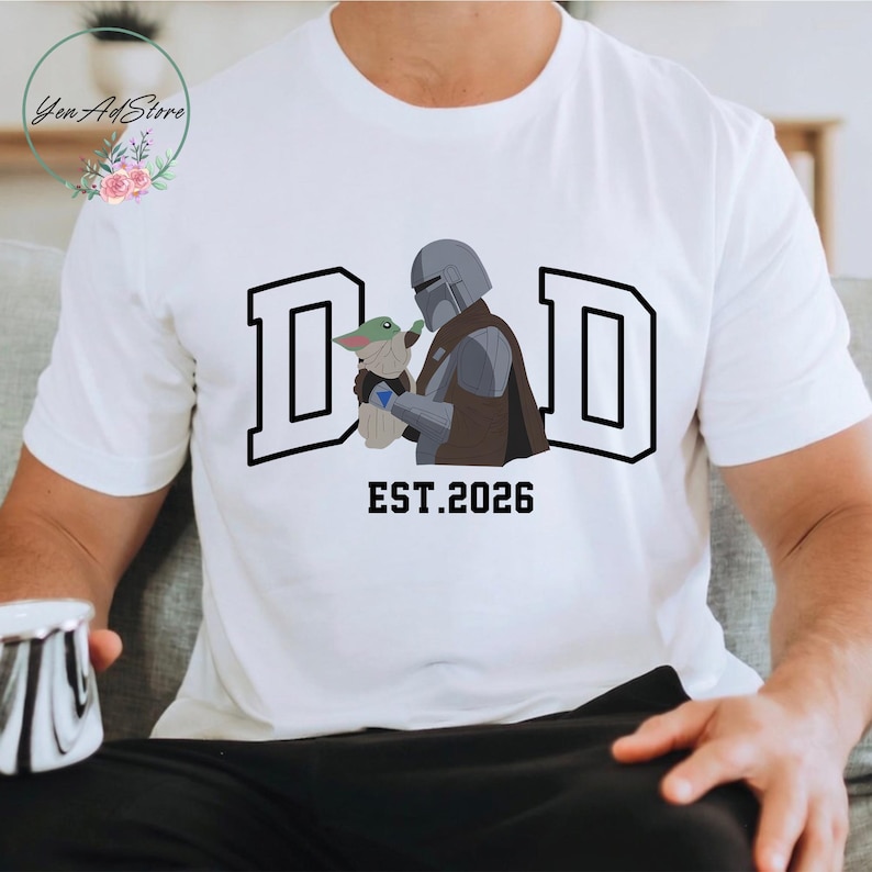 May include: White t-shirt with a graphic of a Mandalorian character holding Grogu, with the word "DAD" in bold black letters. Below the graphic is the text "EST.2026". The shirt is a crew neck and appears to be made of cotton.
