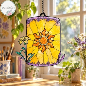 May include: A rectangular stained glass suncatcher featuring a central orange and yellow sun motif with radiating rays. The design includes a small yellow flower on the left and purple accents at the top and bottom edges, all held by a black metal chain.