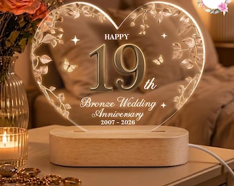 May include: A heart-shaped acrylic plaque on a light wood base, illuminated with the text HAPPY 19th Bronze Wedding Anniversary 2007 - 2026. The design features etched floral vines, leaves, and butterflies surrounding the central number 19.