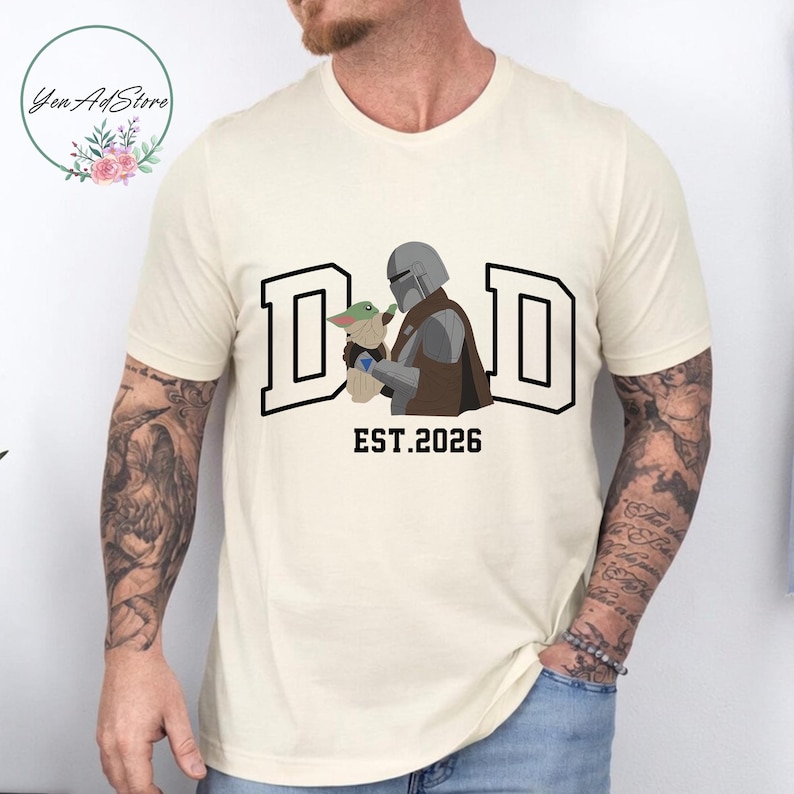 May include: Cream-colored t-shirt featuring the word "DAD" in a bold, black outlined font, with the image of a character holding a small green creature. The text "EST.2026" is below the image. The shirt is a casual wear item.