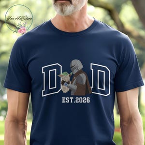 May include: Navy blue t-shirt featuring the word "DAD" in white block letters with a white outline. The graphic includes an illustration of a Mandalorian character holding Grogu, with "EST.2026" below. The shirt is a crew neck.