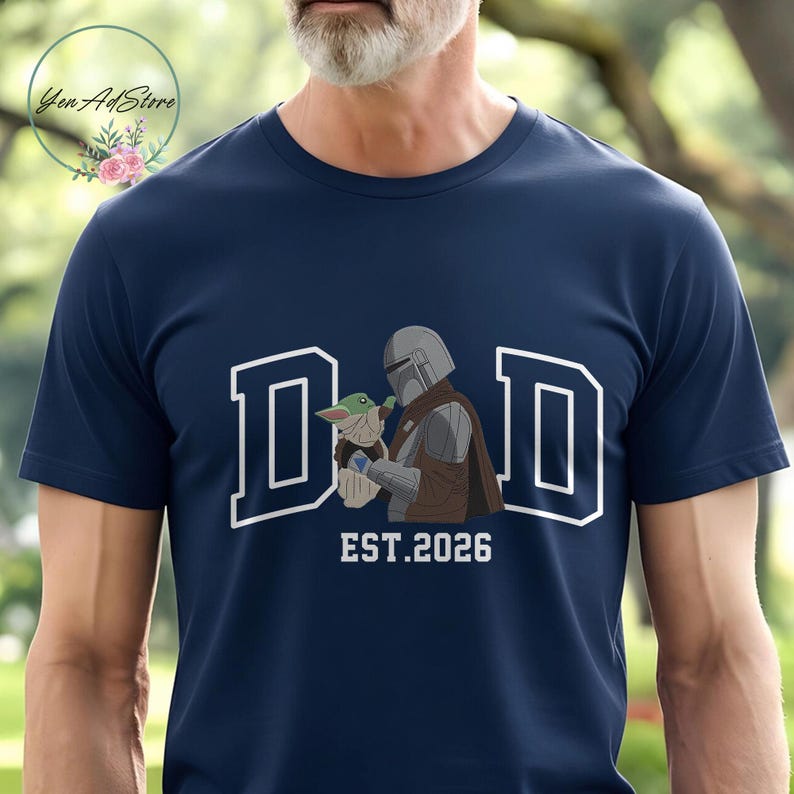 May include: Navy blue t-shirt featuring the word "DAD" in white block letters with a white outline. The graphic includes an illustration of a Mandalorian character holding Grogu, with "EST.2026" below. The shirt is a crew neck.