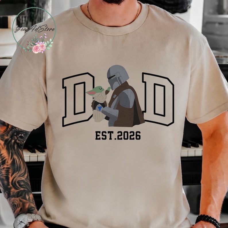 May include: Beige t-shirt with a graphic of a Mandalorian character holding a Grogu figure, with the word "DAD" in bold letters and "EST.2026" below. The design is in black, gray, and brown.