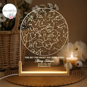 Op de afbeelding: Een rond nachtlampje van acryl met een gegraveerde sterrenkaart van de nachtelijke hemel. De tekst luidt THE SKY THE NIGHT YOU WERE BORN, Marey Johnson, August 8th, 2003, WASHINGTON, DC. Het staat op een rechthoekige houten voet met interne verlichting.