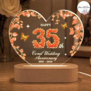 Könnte beinhalten: Eine herzförmige Acrylplatte auf einem hellen Holzsockel mit korallenfarbenen Zweigen, Schmetterlingen und dem Text: HAPPY 35th Coral Wedding Anniversary 1991 - 2026. Das Design enthält leuchtende Elemente und Sternakzente auf der Fläche.