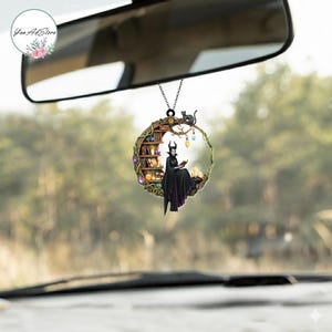 May include: A circular car charm featuring a witch reading a book, with a black cat perched above. The charm has a library and candles inside a decorative frame, hanging from a chain.