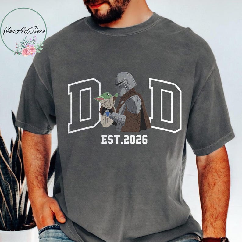 May include: Dark gray t-shirt with a white graphic that says "DAD" in block letters, with the Mandalorian holding Grogu. The text "EST. 2026" is below the graphic. The shirt is a casual style.