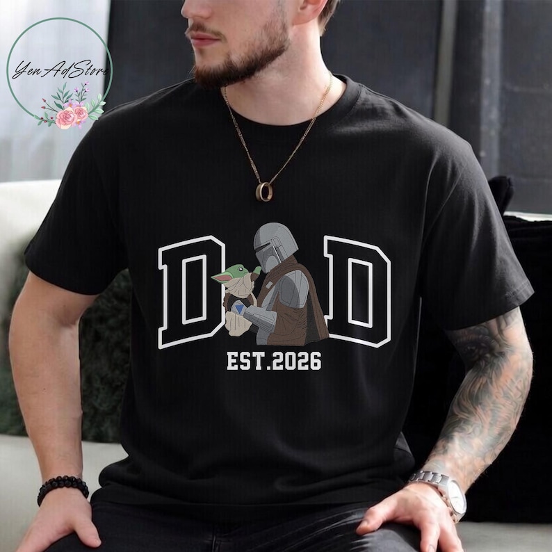 May include: Black t-shirt with a white outlined "DAD" graphic and the text "EST.2026". The graphic features a Mandalorian character holding a small green creature. The shirt is a crew neck.