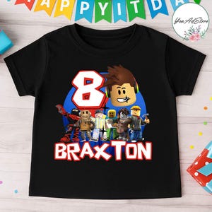 May include: Black t-shirt featuring a colorful graphic design. The design includes the number 8 in blue, Roblox characters, and the name "Braxton" in red. The shirt is likely for a child's birthday.