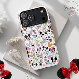 May include: A white phone case featuring a colorful pattern of Disney characters, including Mickey Mouse, Minnie Mouse, Donald Duck, and Goofy. The design includes icons of treats and flowers. The case has a black camera module.