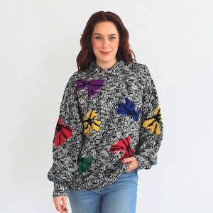 May include: A gray and black sweater with colorful flower appliques. The sweater has a relaxed fit and a high neck. The model is wearing blue jeans and black boots.