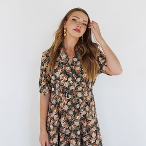 K&ouml;nnte beinhalten: Eine Frau tr&auml;gt ein dunkelgr&uuml;nes und braunes Blumen-Midikleid mit Knopfleiste und G&uuml;rtel. Sie hat lange braune Haare und tr&auml;gt goldene Ohrringe.