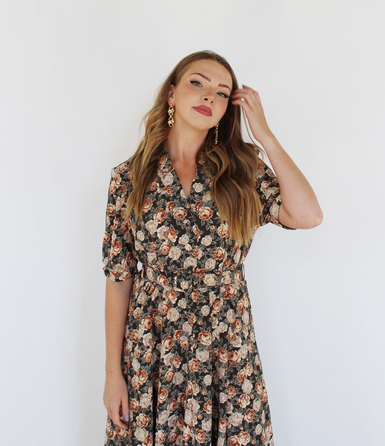 K&ouml;nnte beinhalten: Eine Frau tr&auml;gt ein dunkelgr&uuml;nes und braunes Blumen-Midikleid mit Knopfleiste und G&uuml;rtel. Sie hat lange braune Haare und tr&auml;gt goldene Ohrringe.