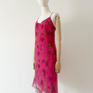 K&ouml;nnte beinhalten: Ein leuchtend pinkfarbenes Slip-Kleid mit einem Rosenbl&uuml;tenmuster. Das Kleid hat d&uuml;nne Tr&auml;ger, einen V-Ausschnitt und einen asymmetrischen Saum. Das Kleid wird auf einer Schaufensterpuppe pr&auml;sentiert.