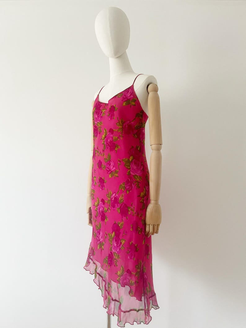 K&ouml;nnte beinhalten: Ein leuchtend pinkfarbenes Slip-Kleid mit einem Rosenbl&uuml;tenmuster. Das Kleid hat d&uuml;nne Tr&auml;ger, einen V-Ausschnitt und einen asymmetrischen Saum. Das Kleid wird auf einer Schaufensterpuppe pr&auml;sentiert.