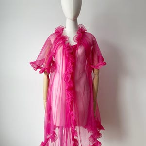 May include: A vibrant fuchsia-pink robe with ruffled detailing, presented on a mannequin. The robe has short sleeves and a knee-length hem. Ruffles adorn the front and edges, giving a delicate, feminine appearance. The garment is crafted from a lightweight, flowing material.