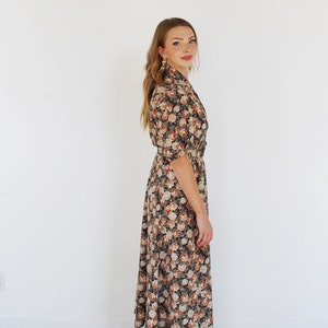 K&ouml;nnte beinhalten: Ein langes, schwarzes Blumenkleid mit einem wei&szlig;en und orangefarbenen Blumendruck. Das Kleid hat einen Kragen, einen G&uuml;rtel in der Taille und Dreiviertel&auml;rmel.