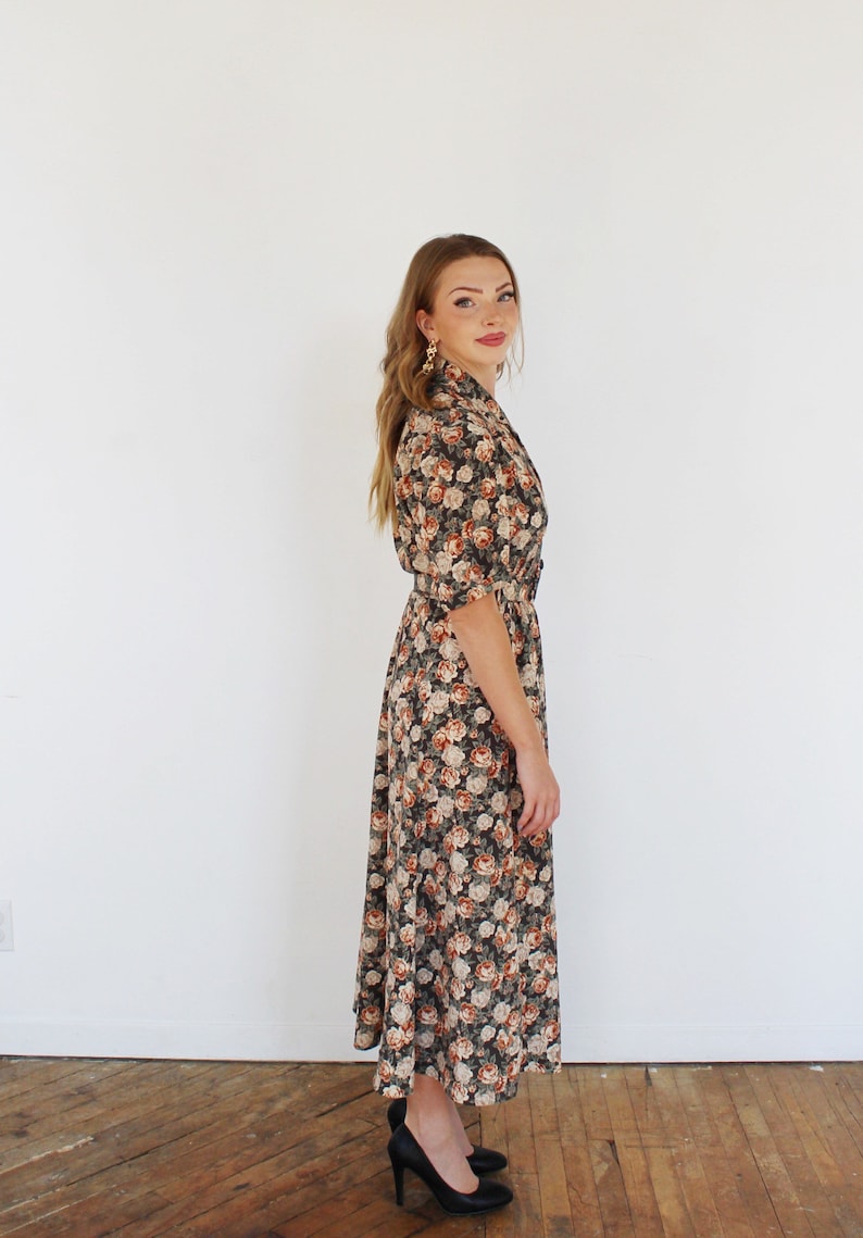 K&ouml;nnte beinhalten: Ein langes, schwarzes Blumenkleid mit einem wei&szlig;en und orangefarbenen Blumendruck. Das Kleid hat einen Kragen, einen G&uuml;rtel in der Taille und Dreiviertel&auml;rmel.