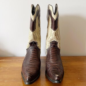 May include: A pair of cowboy boots with a brown, textured leather foot and a cream-colored shaft with decorative stitching. The boots have a pointed toe and a stacked heel, showcasing a classic Western style.