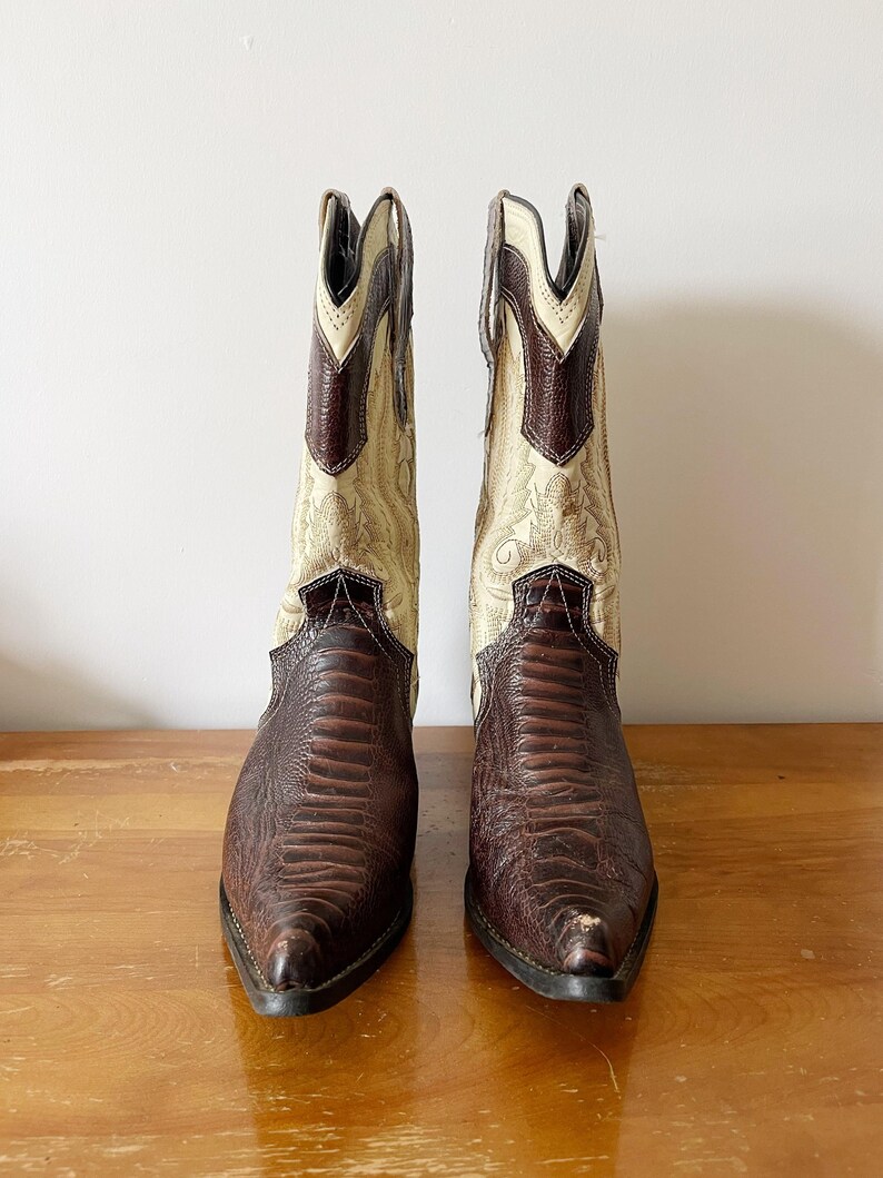 May include: A pair of cowboy boots with a brown, textured leather foot and a cream-colored shaft with decorative stitching. The boots have a pointed toe and a stacked heel, showcasing a classic Western style.